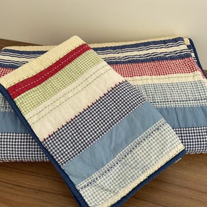 May include: A quilted throw blanket featuring a patchwork design of horizontal stripes in red, light blue, navy blue, green, and cream. The fabric patterns alternate between solid colours, gingham checks, and thin stripes, finished with visible red stitching.