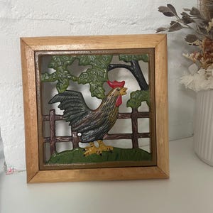 May include: A wooden framed wall decor featuring a rooster in a green field with a brown fence. The rooster is painted in shades of brown, black, red, and yellow.