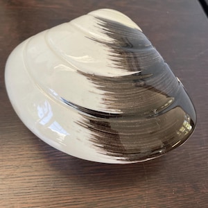May include: A ceramic clam shell-shaped decorative piece. The shell is primarily white with a glossy finish, and features dark brown brushstroke-style accents. The piece is resting on a dark wooden surface.