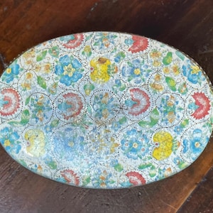 May include: An oval-shaped metal tin featuring a repeating floral pattern in shades of red, blue, yellow, and green against a white background. The lid displays a dense, intricate design of stylised flowers and leaves with a fine dotted texture.