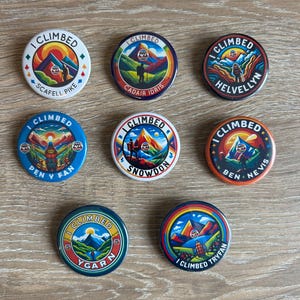May include: Eight circular pin-back badges featuring colourful mountain landscape illustrations. Each badge displays the text I CLIMBED followed by the name of a specific peak: Scafell Pike, Cadair Idris, Helvellyn, Pen y Fan, Snowdon, Ben Nevis, Y Garn, and Tryfan.