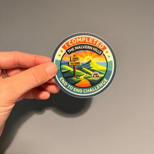 May include: A circular sticker featuring a colorful landscape illustration of the Malvern Hills at sunset. The text reads I COMPLETED THE MALVERN HILLS END TO END CHALLENGE, with a wooden signpost in the center labeled END TO END.