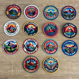 Può includere: Una collezione di toppe rotonde ricamate con disegni di montagne e paesaggi. Ogni toppa presenta testo come "I'VE HIKED" o "I CLIMBED" insieme a nomi di montagne come "Skiddaw" e "Snowdon". Le toppe hanno una varietà di colori.