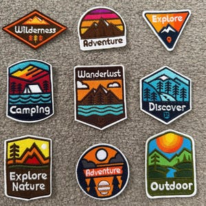 May include: Nine embroidered patches featuring outdoor themes. Designs include mountains, forests, and rivers with text such as Wilderness, Adventure, Explore, Camping, Wanderlust, Discover, Explore Nature, and Outdoor in various shapes and colours.