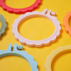 Puede incluir: Una colección de aros de bordado coloridos en varios tonos pastel, incluyendo rosa, azul, amarillo y naranja, con un diseño de borde festoneado. Los aros están dispuestos sobre un fondo amarillo brillante.