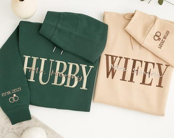 May include: Two matching hoodies, one forest green and one beige. The green hoodie says "Hubby" with "Mr. Henry" underneath, and the date "07.18.2021" with rings on the sleeve. The beige hoodie says "Wifey" with "Mrs. Smith" underneath, and the date "07.18.2021" with rings on the sleeve.