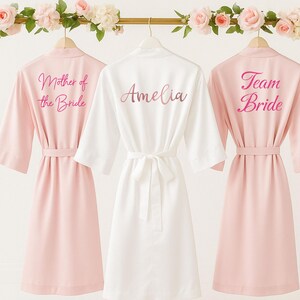 Könnte beinhalten: Drei Brautroben hängen an einem goldenen Gestell, das mit rosa Rosen geschmückt ist. Zwei rosa Roben tragen die Aufschriften "Mother of the Bride" und "Team Bride". Die mittlere Robe ist weiß mit dem Namen "Amelia".