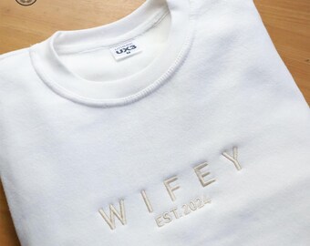 May include: White sweatshirt with the word "WIFEY" embroidered in a light beige thread, along with "EST. 2024". The crew neck sweatshirt is folded neatly. The brand label is visible on the inside of the collar.