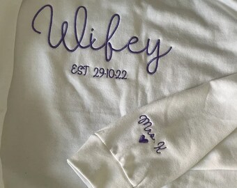 May include: A white sweatshirt with purple embroidered text that reads "Wifey" above "EST 29-10-22". The sleeve features "Mrs. K" and a heart. The sweatshirt is a casual garment.