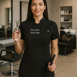 May include: A black salon uniform with short sleeves and a mandarin collar. The uniform has a button detail and the text "Your Own Text/Logo" on the front. The person is holding scissors and a comb.