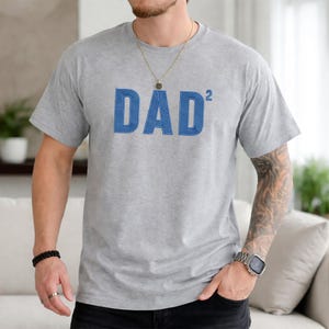 May include: A light gray t-shirt with the word "DAD²" in blue, block letters. The shirt has short sleeves and a crew neck. The text is centered on the chest.