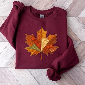 May include: A burgundy sweatshirt with a detailed maple leaf design. The leaf is made from various fabric patterns in orange, yellow, and green, creating a patchwork effect. The sweatshirt has long sleeves and a crew neck.