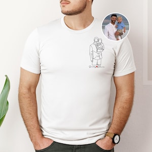 以下が含まれることがあります： 白いTシャツ。ミニマリストな線画で子供を抱く人物が描かれ、赤で「Daddy & [heart] Oliver」の文字。男性と子供の丸い写真が見える。Tシャツは人が着ています。