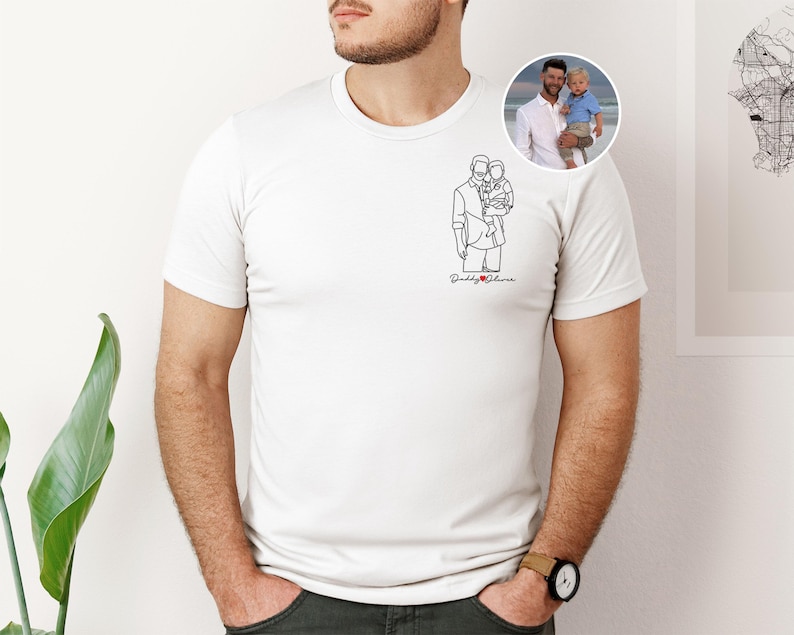 以下が含まれることがあります： 白いTシャツ。ミニマリストな線画で子供を抱く人物が描かれ、赤で「Daddy & [heart] Oliver」の文字。男性と子供の丸い写真が見える。Tシャツは人が着ています。
