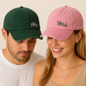 Puede incluir: Gorra de béisbol verde oscuro con la palabra "Mr." bordada y gorra de béisbol rosa con la palabra "Mrs." bordada. Llevadas por una pareja.