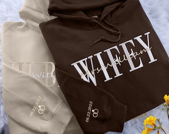 May include: A dark brown hoodie with white embroidered text reading WIFEY and Mrs. William, and a beige hoodie with matching embroidery reading HUBBY and Mr. William. Both sleeves feature the date 06.22.2023 and two interlocking ring icons.