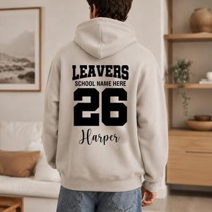 May include: A light beige hooded sweatshirt featuring black text on the back that reads LEAVERS, SCHOOL NAME HERE, the number 26, and the name Harper in a script font. The hoodie has long sleeves, a hood, and a ribbed hem and cuffs.