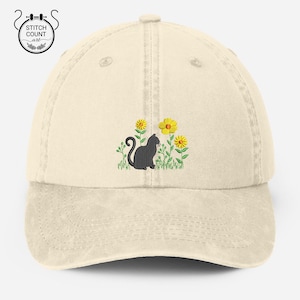 May include: A beige baseball cap with an embroidered black cat sitting in a field of yellow flowers.