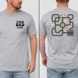 Puede incluir: Camiseta gris claro con el texto "TIRED DAD CLUB" en la parte delantera. La parte posterior de la camiseta presenta un diseño de mapa de carreteras colorido con casas, árboles y coches. La camiseta es de material suave.