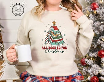 以下が含まれることがあります： 積み重ねた本で形作られたクリスマスツリーのグラフィックが描かれたベージュのクルーネックトレーナー。ツリーにはイルミネーションライトと星の飾りが付いています。ツリーの下には赤と緑で「ALL BOOKED FOR Christmas」という文字がプリントされています。