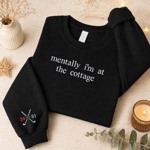 May include: Black sweatshirt with the text "mentally I'm at the cottage" in white. The left sleeve has embroidered golf clubs and the numbers 24 and 81. The sweatshirt is a crew neck style.