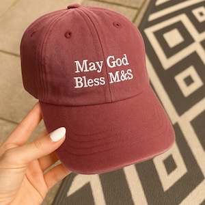 Pode incluir: Boné de beisebol bordô com a inscrição bordada em branco "May God Bless M&S". O boné tem uma viseira curva e um botão coberto de tecido. O chapéu é feito de um material macio e texturizado.