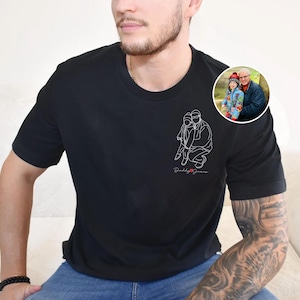 以下が含まれることがあります： 白線画で人物と子供を描いた黒のクルーネックTシャツ。「Daddy Jeans」の文字が筆記体で描かれています。人物と子供の小さな円形画像が右肩にあります。