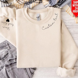 May include: A beige sweatshirt with the text "Custom text" on the right shoulder and the text "Forever you" with a heart on the left sleeve.