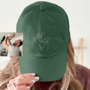 Könnte beinhalten: Eine grüne Baseballkappe mit einer weißen Umrandungsstickerei einer Person, die ein Kind hält. Die Stickerei befindet sich auf der Vorderseite der Kappe.