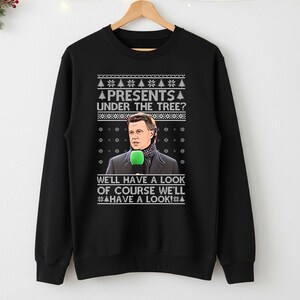 May include: Black crew neck sweatshirt with a festive design. The graphic features a man holding a microphone, with the text "PRESENTS UNDER THE TREE? WE'LL HAVE A LOOK OF COURSE WE'LL HAVE A LOOK!" in white.