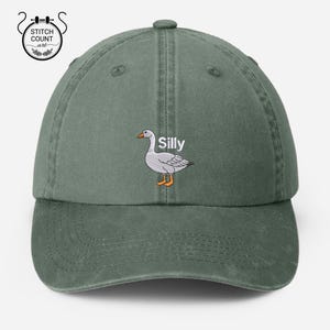 May include: A green, adjustable, cotton twill baseball cap with an embroidered white goose and the word "Silly" in white text.