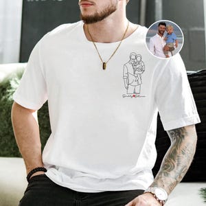 以下が含まれることがあります： 白いTシャツ。線画で子供を抱いている人物が描かれており、黒い筆記体で「Daddy & Olivia」と書かれています。子供を抱いている人物の小さな丸い写真が右上隅にあります。その人物は金のネックレスをしています。