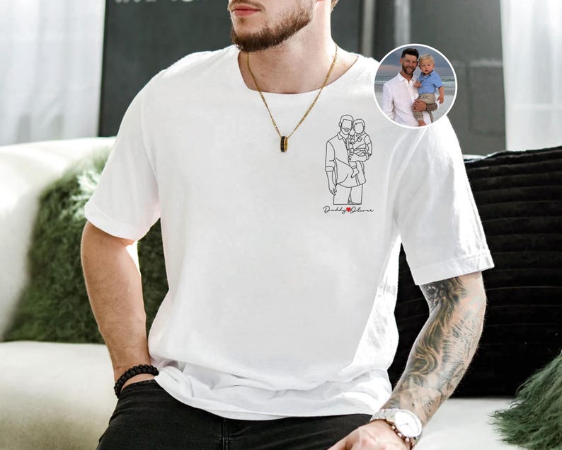 以下が含まれることがあります： 白いTシャツ。線画で子供を抱いている人物が描かれており、黒い筆記体で「Daddy & Olivia」と書かれています。子供を抱いている人物の小さな丸い写真が右上隅にあります。その人物は金のネックレスをしています。