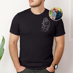 以下が含まれることがあります： 黒いTシャツ。白い線画で子供を抱きしめる人のイラスト、筆記体で「Daddy James」の文字。男性と子供の小さな丸い写真が見える。Tシャツは人が着ています。