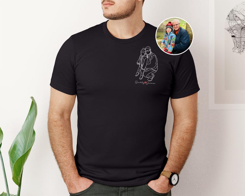 以下が含まれることがあります： 黒いTシャツ。白い線画で子供を抱きしめる人のイラスト、筆記体で「Daddy James」の文字。男性と子供の小さな丸い写真が見える。Tシャツは人が着ています。