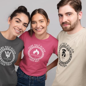 May include: Three people wearing gray, pink, and beige t-shirts with Camp Half-Blood logos. The logos feature different Greek god symbols and cabin numbers. The text on the shirts reads "Camp Half-Blood", "Poseidon", "Cabin Three", "Hephaestus", "Cabin Nine", "Ares", and "Cabin Five".
