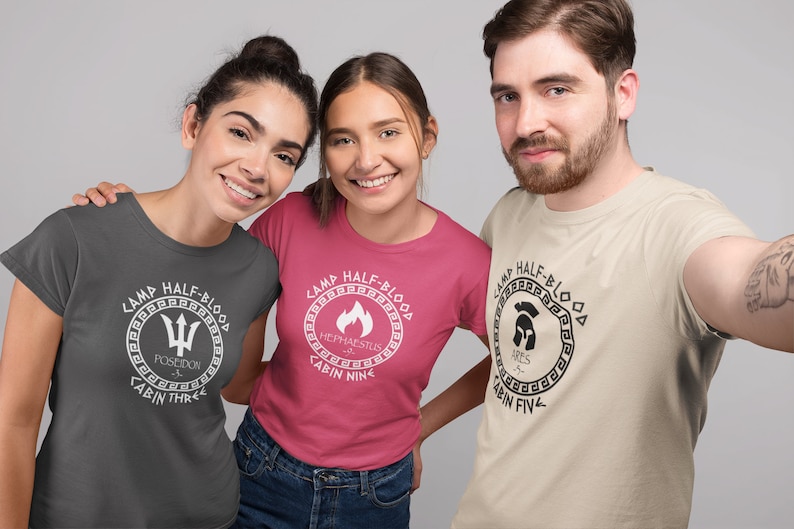 May include: Three people wearing gray, pink, and beige t-shirts with Camp Half-Blood logos. The logos feature different Greek god symbols and cabin numbers. The text on the shirts reads "Camp Half-Blood", "Poseidon", "Cabin Three", "Hephaestus", "Cabin Nine", "Ares", and "Cabin Five".