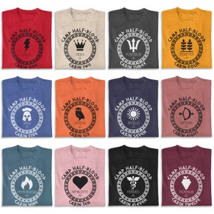 May include: Twelve t-shirts with different designs featuring the Greek god or goddess name and cabin number from the Camp Half-Blood series. The shirts are in various colors, including red, blue, orange, yellow, pink, and black.