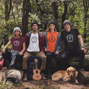May include: Four people wearing Camp Half-Blood t-shirts with different cabin logos sit on a log in a forest. Two dogs are lying in front of them. One person is holding a ukulele.