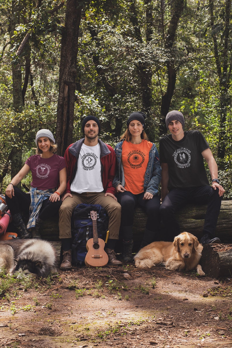 May include: Four people wearing Camp Half-Blood t-shirts with different cabin logos sit on a log in a forest. Two dogs are lying in front of them. One person is holding a ukulele.