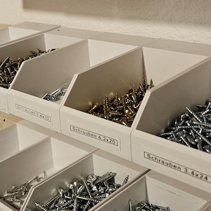 Könnte beinhalten: Weiße Kunststoff-Aufbewahrungsbehälter mit verschiedenen Größen von Schrauben und Muttern. Die Behälter sind mit dem Text "Schrauben 3, 4x24" und "Schrauben 4, 2x20" beschriftet.