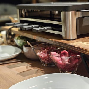 Könnte beinhalten: Ein Raclette-Grill aus Edelstahl mit schwarzer Oberseite und Holzregalen. Unter dem Grill befinden sich Glasschalen mit gewürfeltem Fleisch. Weiße Teller befinden sich im Vordergrund, was auf eine Essumgebung hindeutet.