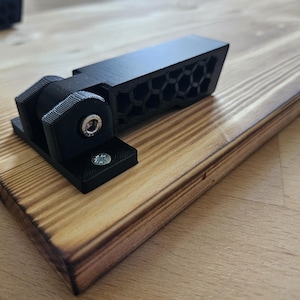 Könnte beinhalten: Ein schwarzer, 3D-gedruckter Halter mit Wabenmuster, montiert auf einer gebeizten Holzplanke. Die Halterung hat einen Schwenkmechanismus und ist mit Schrauben befestigt. Das Holz hat eine natürliche Maserung und eine warme, braune Farbe.
