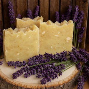 May include: Three rectangular bars of handmade soap with a textured top, in a light yellow color, are displayed with sprigs of purple lavender. The soap is on a wooden slice, with a wooden background.