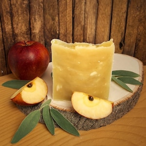 Puede incluir: Una barra rectangular de jabón amarillo con la parte superior rugosa, exhibida con rodajas de manzana, una manzana roja entera y hojas verdes sobre una rodaja de madera. El fondo es una pared de madera.