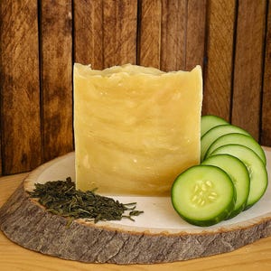 May include: A large, pale yellow bar of soap with a textured surface, presented on a wooden slab. Sliced green cucumbers and dried green tea leaves are arranged around the soap, suggesting natural ingredients.