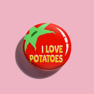 May include: A round pinback button featuring a bright red background with a stylised green tomato stem graphic at the top. The text I LOVE POTATOES is printed in bold yellow block letters across the centre of the circular surface.