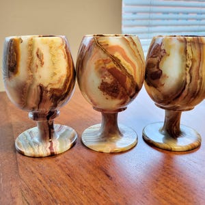Peut inclure: Trois verres à vin décoratifs en pierre naturelle. Les verres ont une forme arrondie et une courte tige avec une base circulaire. La pierre présente un mélange de couleurs marron, beige et crème.