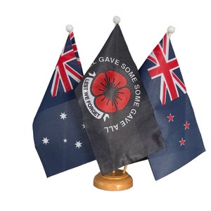 May include: Three small flags on a wooden stand. The centre flag is black with a red poppy and the text "LEST WE FORGET SOME GAVE ALL SOME GAVE SOME". The flags on either side are the Australian and New Zealand flags.