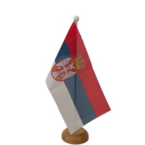 May include: A small desk flag of Serbia. The flag is red, white, and blue with the coat of arms of Serbia in the centre. The flag is mounted on a wooden base.